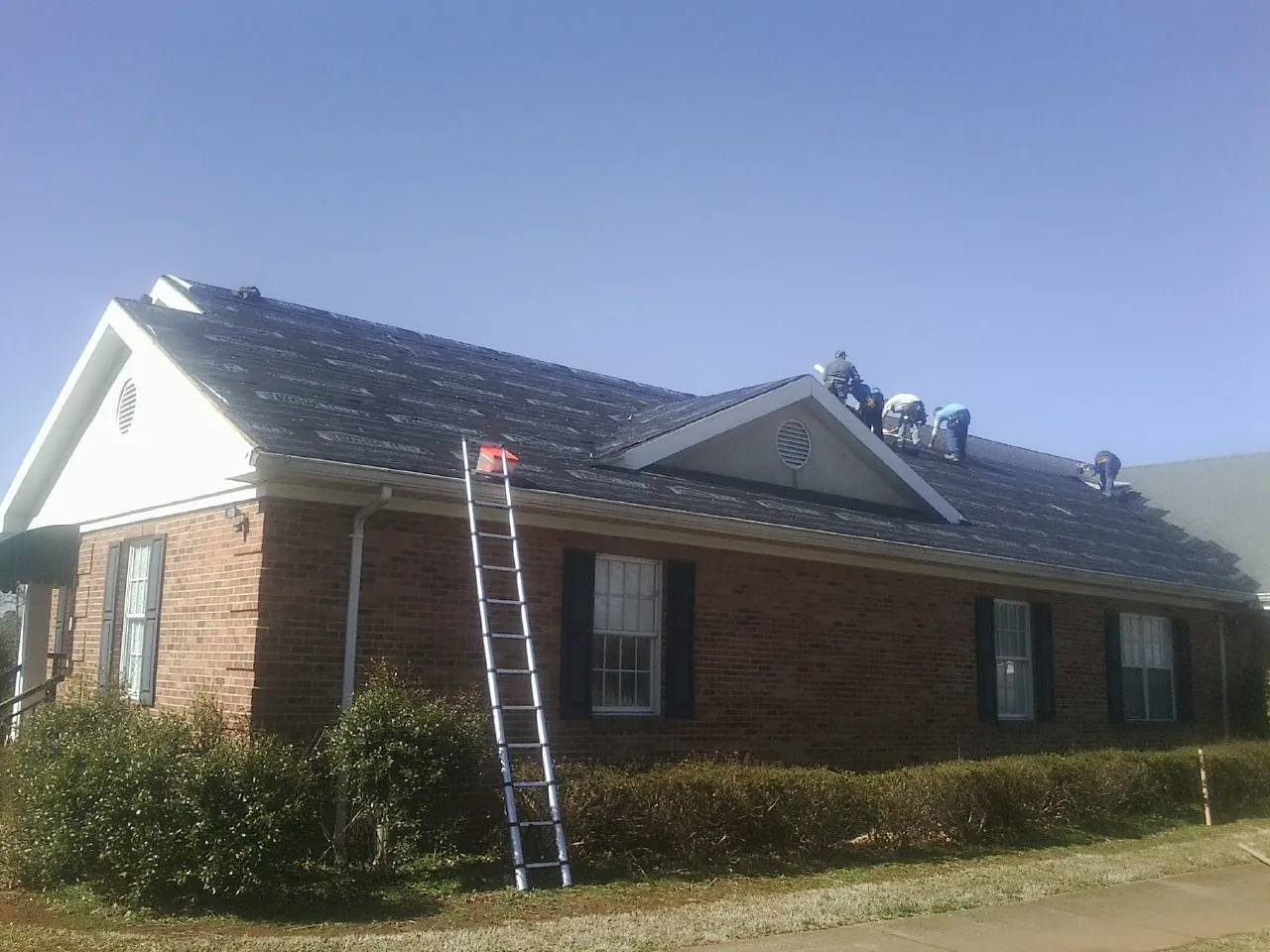 Crew tearing shingles off a brick ranch roof with a ladder set — Emergency Roof Repair work by our crew in Ihlen